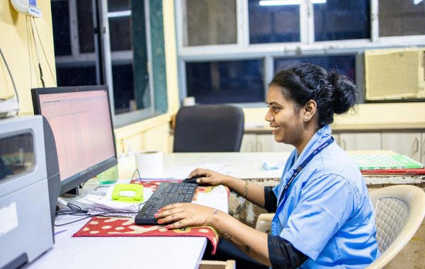 A woman in a blue uniform and a lanyard sits at a desk with a computer, keyboard and mouse